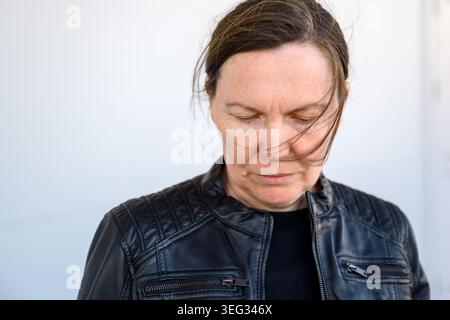 Donna di mezza età con una giacca in pelle nera che guarda in basso con capelli soffiati dal vento e una messa a fuoco selettiva Foto Stock