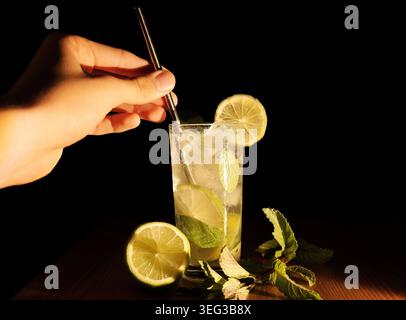 Mescolando a mano un classico cocktail mojito con fette di lime fresche, foglie aromatiche di menta e ghiaccio, presentato in un alto bicchiere contro un backgroun scuro Foto Stock