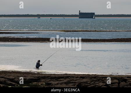 Ile-d'Aix, Francia - 27 agosto 2018: Un pescatore solitario in acque poco profonde getta una lunga canna con il famoso Fort Boyard in piedi all'orizzonte tra oy Foto Stock