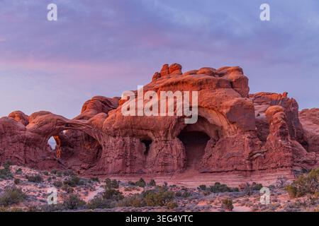 Formazioni di roccia rossa con archi naturali brillano nella calda luce del tramonto contro un cielo viola serale. Foto Stock