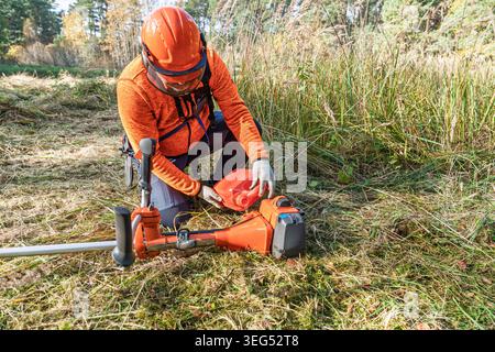 Man rifornisce il decespugliatore sul campo, indossando dispositivi di sicurezza per lavori di manutenzione paesaggistica Foto Stock