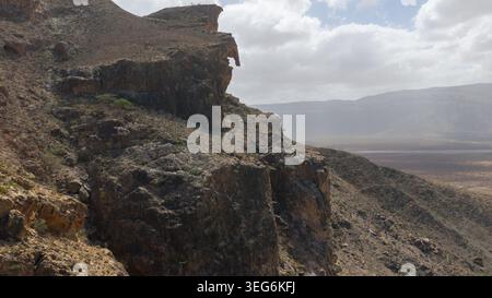 Scogliera rocciosa che domina l'arida valle sotto il cielo nuvoloso, aspro paesaggio di montagna con formazioni rocciose a strati e colline lontane in luce nebbiosa. Sok Foto Stock