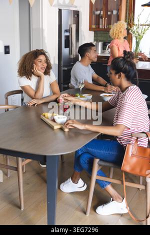 Amici diversi che si riuniscono al tavolo della cucina di casa, donna in maglietta a righe che cerca i cracker. Gruppo, informale, soleggiato, candid, cena, rustico, in legno Foto Stock