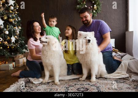 Famiglia felice con il cane samoyed husky seduto vicino al bellissimo albero di Natale a casa. Foto Stock