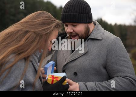 Giovani coppie all'aperto in autunno scambiandosi un regalo a sorpresa, sorridendo e abbracciando in un sincero momento di gioia, amore e festività insieme durante il freddo Foto Stock