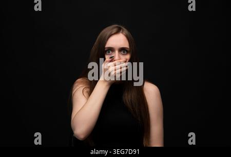 Giovane donna che si copre la bocca con la mano, esprimendo shock e paura con gli occhi spalancati su uno sfondo scuro Foto Stock