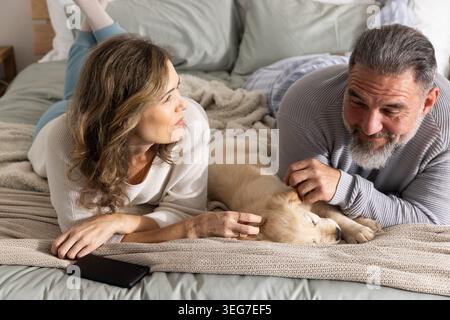 Coppia sdraiata sul letto con il lancio, uomo che accarezza il cucciolo dorato, donna che guarda al telefono dai cuscini Foto Stock