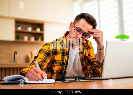 Con un'espressione pensiva, un uomo lavora sul suo portatile e scrive in blocco note, sembrando analizzare le informazioni o risolvere un problema online. Foto Stock