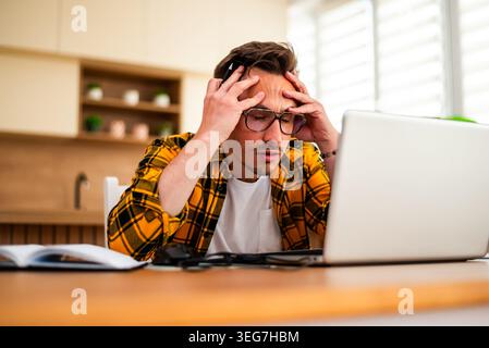 Un giovane stressato lavora sul suo portatile tenendo una penna, appoggiando la testa in mano, mostrando segni di frustrazione o stanchezza mentale. Foto Stock