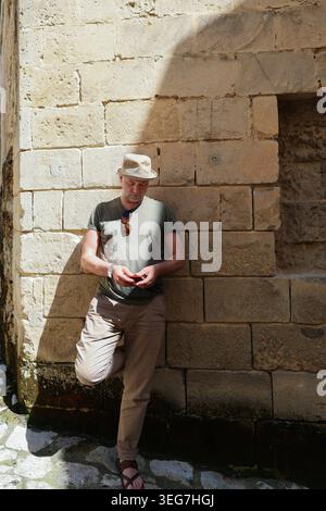 Elegante uomo di mezza età che usa lo smartphone mentre si trova vicino a un muro di pietra storico nella soleggiata strada della città vecchia europea. Foto Stock