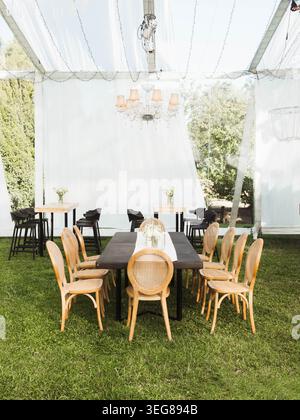 La tenda bianca protegge da un elegante ambiente da pranzo all'aperto. Il tavolo in legno con sedie intrecciate invita gli ospiti a riunirsi. Il lampadario si illumina delicatamente sopra il centro Foto Stock
