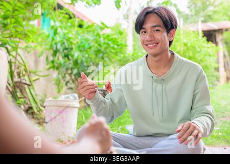 Un ritratto orizzontale di un allegro giovane asiatico della regione di Isan in Thailandia seduto all'aperto a gambe incrociate. Guarda direttamente il Foto Stock