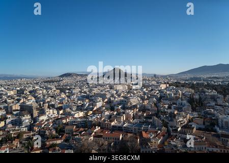 Un'ampia vista aerea cattura il denso paesaggio urbano di Atene in Grecia sotto un cielo azzurro, con il colle di Licabetto e le montagne circostanti Foto Stock