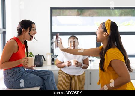 Diverse amiche che ridono, puntano lo smartphone e tengono in mano tazze in ceramica nella cucina moderna Foto Stock