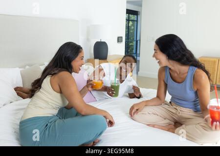 Diverse amiche che si siedono e parlano sul letto in camera da letto tenendo delle bevande indossando abiti casual Foto Stock