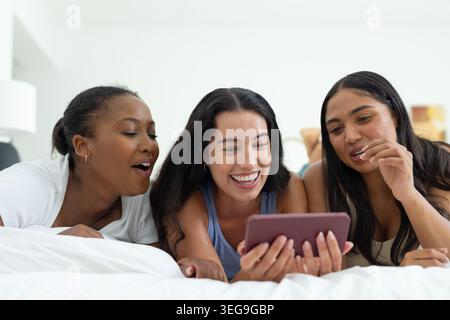 Diverse amiche sul letto, che si appoggiano e guardano un tablet, indossano canotte Foto Stock