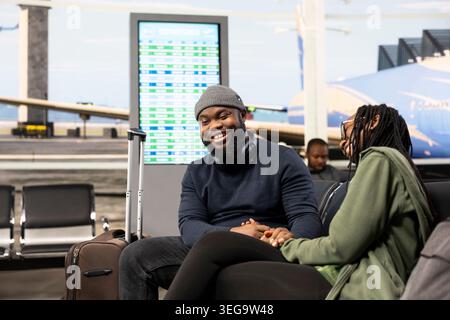 Simpatica coppia nera che si coccola e ride aspettando l'aereo, si gode una vacanza al terminal dell'aeroporto e si sente emozionata. I turisti amano parlare insieme di visite turistiche e tempo libero. Foto Stock