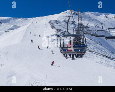 Una scena invernale a la Thuile, che mostra una seggiovia per quattro persone che sale su una pista innevata, con gli sciatori che scendono lungo la pista ben curata sotto un cielo limpido Foto Stock
