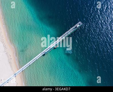 Questa splendida fotografia aerea cattura un lungo molo di legno che si estende nelle limpide acque blu dell'oceano, circondato da una spiaggia di sabbia su un lato Foto Stock