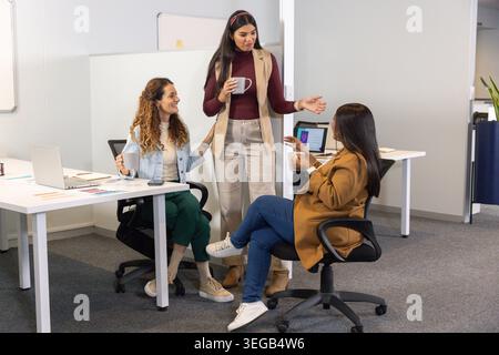 Diverse collaboratrici che chiacchierano e gestivano le scrivanie di un ufficio moderno con computer portatili e tazze Foto Stock