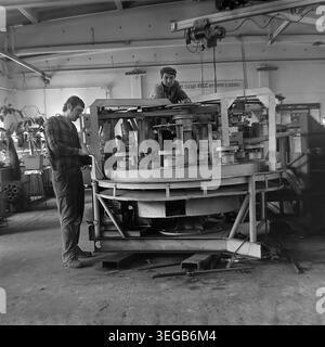 Sloviansk, 1981, AIZ (impianto di isolamento di armature). Due meccanici maschi sono profondamente concentrati sulla riparazione o l'assemblaggio di una macchina industriale circolare grande e complessa in officina. Un uomo sta in piedi a esaminare una scatola di controllo, mentre l'altro si appoggia sul meccanismo rotante. Una storica fotografia d'archivio in bianco e nero che cattura l'atmosfera autentica dell'epoca, il processo di manutenzione tecnica altamente qualificato e la vita quotidiana dei lavoratori. Uno sguardo dettagliato sul passato, sull'industria sovietica e sul pacifico Donbas. Foto Stock