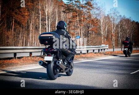 Motociclista con attrezzatura protettiva, casco, motocicletta con valigia su strada asfaltata. Segue un altro motociclista. Sfondo della foresta, cielo limpido. TR Foto Stock