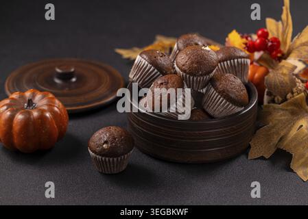 Deliziosi muffin al cioccolato riempiono un recipiente in ceramica, disposti ad arte su uno sfondo scuro tra foglie decorative autunnali, una piccola zucca e un berro rosso Foto Stock