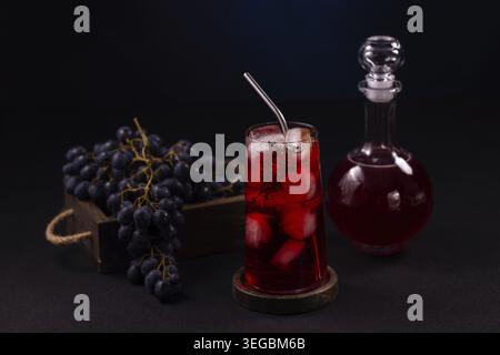 Cocktail di succo d'uva rossa in un bicchiere alto con ghiaccio e paglia metallica accanto ad un mucchio di uva scura e ad una caraffa di vetro su un suggestivo sfondo scuro, Foto Stock
