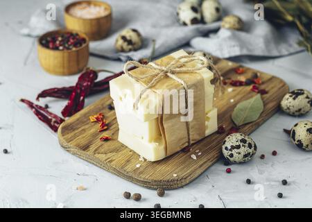 Fette di formaggio Maasdam, avvolte in carta marrone e spago, disposte su un asse di legno con uova di quaglia, peperoncino essiccato e spezie Foto Stock