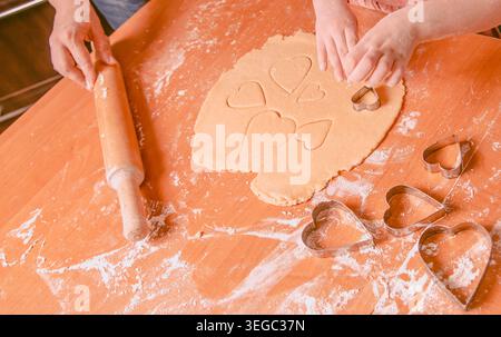 Due bambini usano le forbici a forma di cuore per modellare l'impasto su un tavolo di legno, con farina sparsi intorno e un mattarello nelle vicinanze Foto Stock