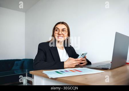 Donna seduta alla scrivania che tiene lo smartphone e sorride. Affari. Lavoro d'ufficio. Bella donna professionale sicura di sé e rilassata. Beauty portra Foto Stock