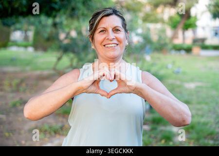 Donna anziana in piedi all'aperto, sorridendo e gestendo un cuore con le mani, simboleggiando l'amore, la salute e il benessere Foto Stock