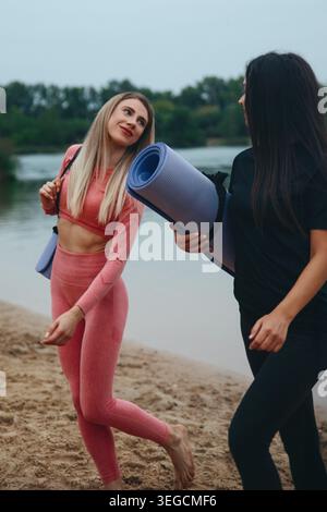 Due donne camminano lungo la riva sabbiosa del lago portando con sé stuoie per lo yoga, sorridendo e chiacchierando sul loro percorso di fitness in una splendida giornata. Foto Stock