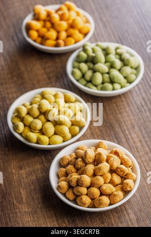 Arachidi rivestite assortite servite in piccole ciotole bianche su un tavolo di legno, caratterizzate da croccanti e aromatizzati in giallo, arancione e verde. Ciotole colorate ideali per feste, antipasti o spuntini informali. Foto Stock