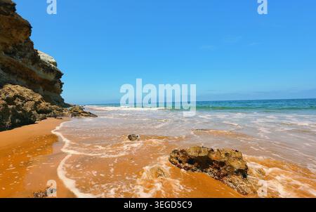 Spiaggia di sabbia dorata con onde che si infrangono dolcemente contro aspre scogliere sotto un cielo azzurro Foto Stock
