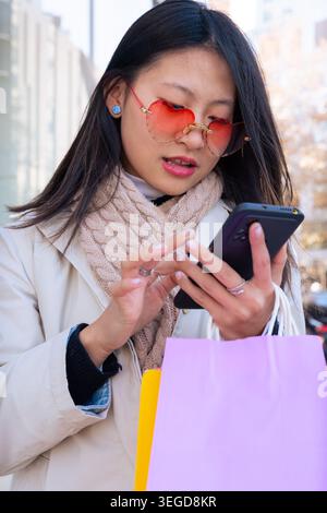 Ritratto verticale giovane donna con borse della spesa che usa lo smartphone per strada Foto Stock