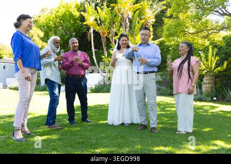 Sposo spumante, sposa che indossa un abito da sposa con flauto, ospiti che festeggiano sul prato Foto Stock