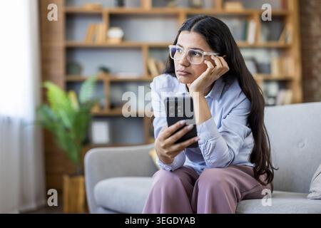 Giovane donna che indossa gli occhiali da vista e prova noia o solitudine mentre siede su un comodo divano a casa, tenendo e guardando distrattamente il suo smartphone durante il tempo libero in casa Foto Stock