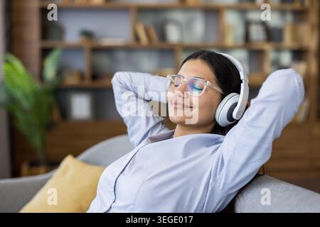 Giovane donna con gli occhiali che si rilassa su un divano, le mani dietro la testa, sorride mentre ascolta la musica con le cuffie, godendosi momenti di relax tranquilli e confortevoli a casa Foto Stock