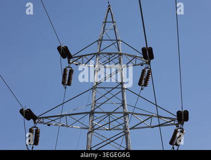 ripresa ad angolo basso di una torre di trasmissione a reticolo in acciaio che trasporta linee di alimentazione ad alta tensione su un chiaro sfondo blu del cielo Foto Stock