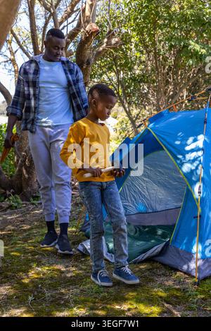 Padre e ragazzo afroamericano che lanciano tenda blu nei boschi, ragazzo che tiene in mano la mazzetta, paletto della tenda Foto Stock