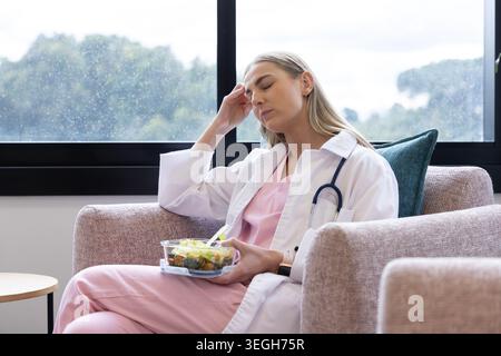 Medico donna adulta seduta nella sala con scrub rosa con insalata e stetoscopio che sfrega il tempio Foto Stock