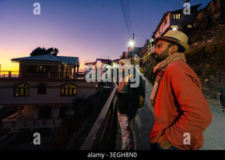 Due persone in piedi su un sentiero collinare che si affaccia sulle case tradizionali durante un colorato tramonto a Chakrata, Uttarakhand, India. Foto Stock