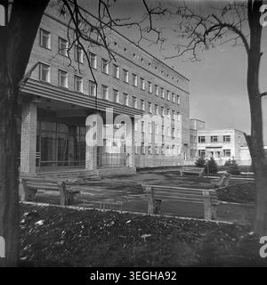 Sloviansk, fine anni '1970 - inizio anni '1980, ospedale pediatrico locale. Una prospettiva angolata si concentra sull'ingresso principale e sull'ampio esterno in mattoni dell'istituto medico. L'edificio presenta un grande baldacchino sostenuto sopra le porte di vetro e numerose finestre uniformi che riflettono la luce del sole. Il cortile anteriore comprende sentieri pavimentati, diverse panchine di legno vuote per i visitatori e piccoli alberi decorativi di conifere piantati nel terreno scuro. Questa foto d'archivio documenta splendidamente i servizi pubblici urbani e un tranquillo assaggio di pace nel Donbas. Foto Stock