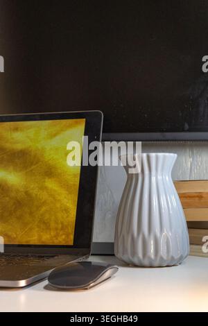 Un notebook aperto con schermo dorato, un mouse wireless e un vaso in ceramica scanalato si trovano su una scrivania bianca. Foto Stock