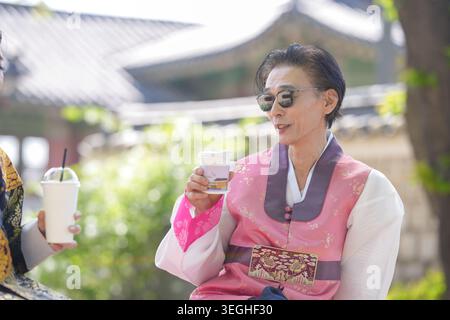 Una donna con un tradizionale abbigliamento coreano regge una tazza di caffè. Indossa gli occhiali da sole, sorride dolcemente. La luce soffusa del sole si illumina attraverso gli alberi dietro di lei. Sho. Sfondo Foto Stock