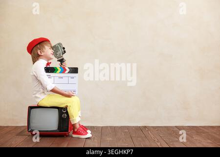 Simpatico ragazzo che tiene in mano una macchina fotografica vintage e applaude sullo sfondo grunge della parete. Ragazzo seduto sulla vecchia TV. Bambino che gioca a casa Foto Stock