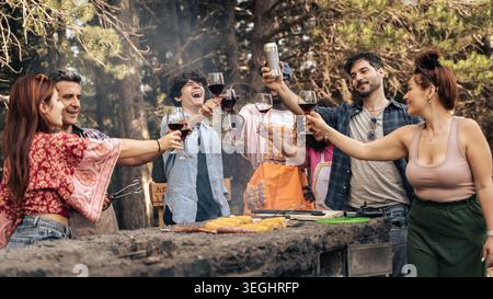 Un gruppo di amici sta intorno a un grill, alzando le bevande e ridendo. Stanno cucinando il cibo su una fiamma libera in un ambiente forestale durante l'Summe Foto Stock