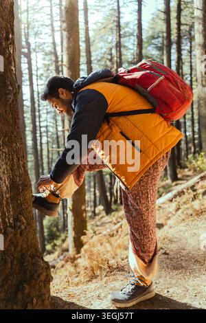 Un giovane uomo che indossa un gilet giallo e uno zaino rosso lega gli stivali da trekking su un sentiero sterrato in una pineta a Chakrata, Uttarakhand, India. Foto Stock