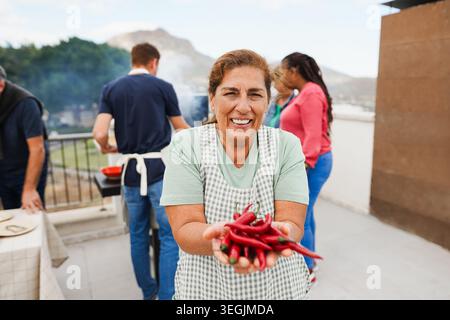 Donna latina anziana che tiene in mano il peperoncino fresco mentre cucina il pranzo al barbecue sullo sfondo - persone multigenerazionali che si divertono insieme durante il fine settimana Foto Stock